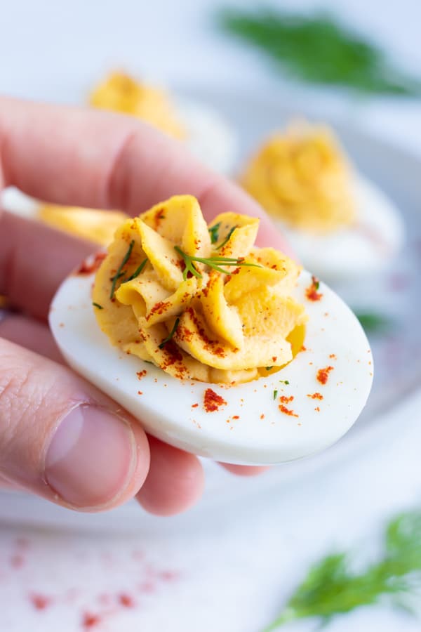 A hand picking up a hard-boiled egg with a creamy yolk filling with a fresh dill sprig on top.