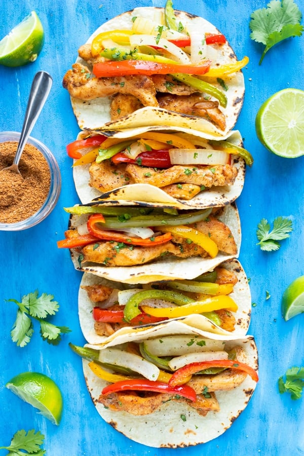 Chicken fajita tacos in a row with bell peppers and onions next to taco seasoning and limes.