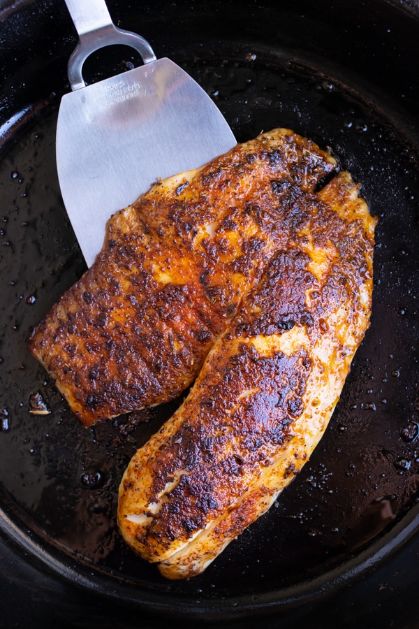 A spatula picking up a filet of blackened tilapia in a cast iron skillet with blackened seasoning.