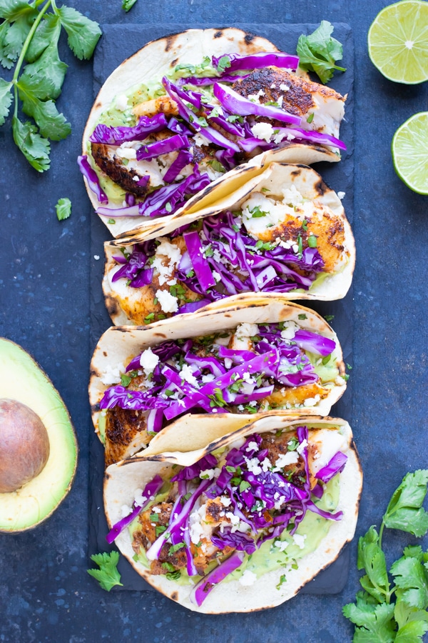 Four blackened fish tacos with avocado sauce and cabbage on a black plate.