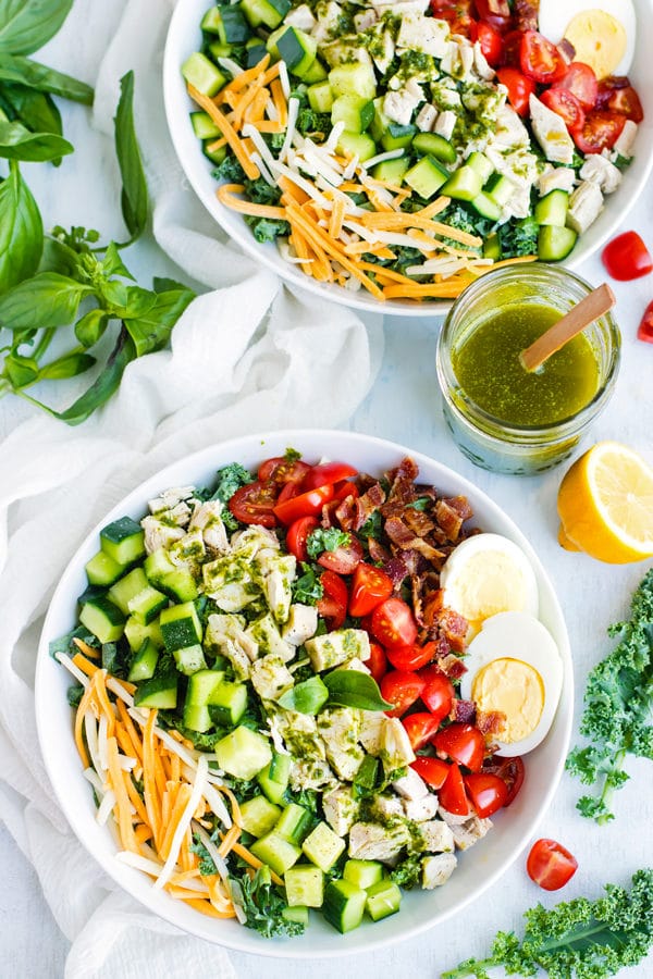 Two white bowls full of chicken cobb kale salad with a drink in the middle.