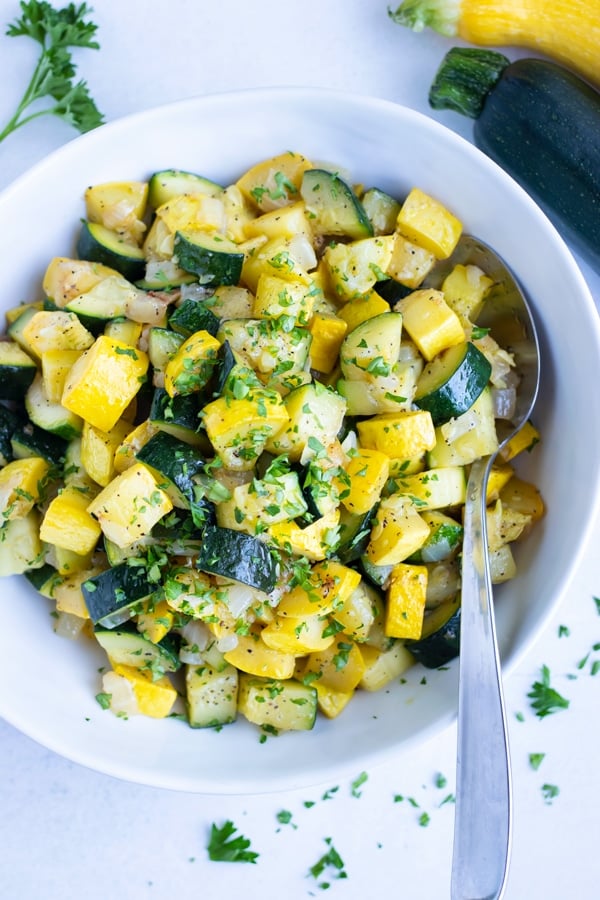 Sautéed zucchini and yellow summer squash in a white bowl with a serving spoon.