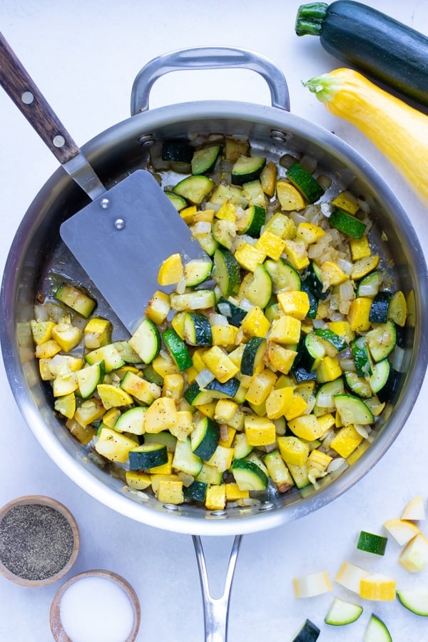 Sautéed zucchini and yellow summer squash in a stainless steel skillet with a spatula.