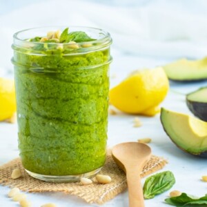 Gluten-free vegan pesto recipe in a glass jar with avocados and lemons on the side.