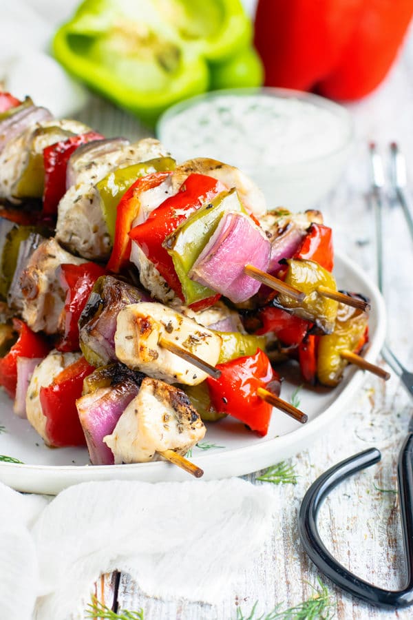 Side picture of a pile of Grilled Greek Chicken Shish Kabobs with sauce in the background.