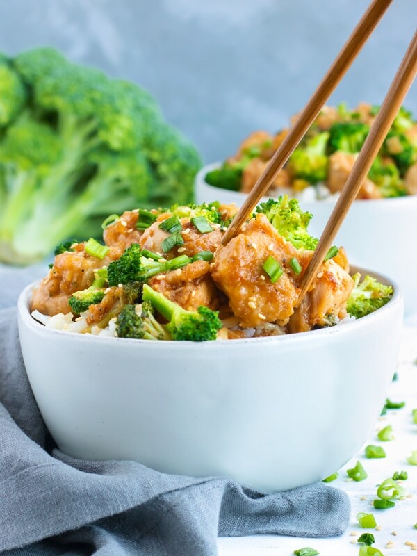 Two white bowls of Honey Sesame Chicken and Broccoli Stir-Fry with chopsticks holding a piece of chicken.