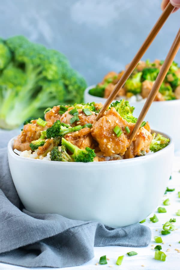 Two white bowls of Honey Sesame Chicken and Broccoli Stir-Fry with chopsticks holding a piece of chicken.