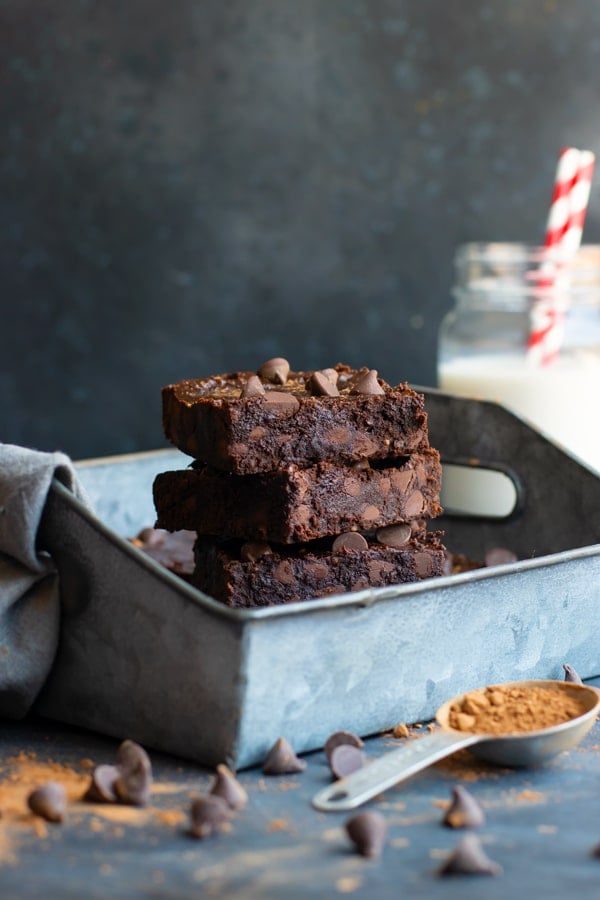 Stacked paleo brownies in a silver tray with a measuring spoon in front.