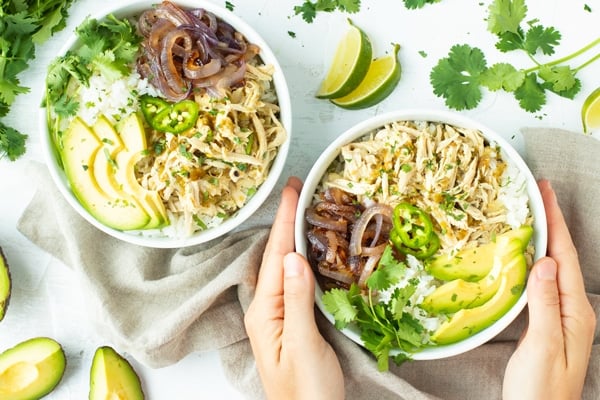 A hand touching a white bowl filled with crockpot shredded chicken.