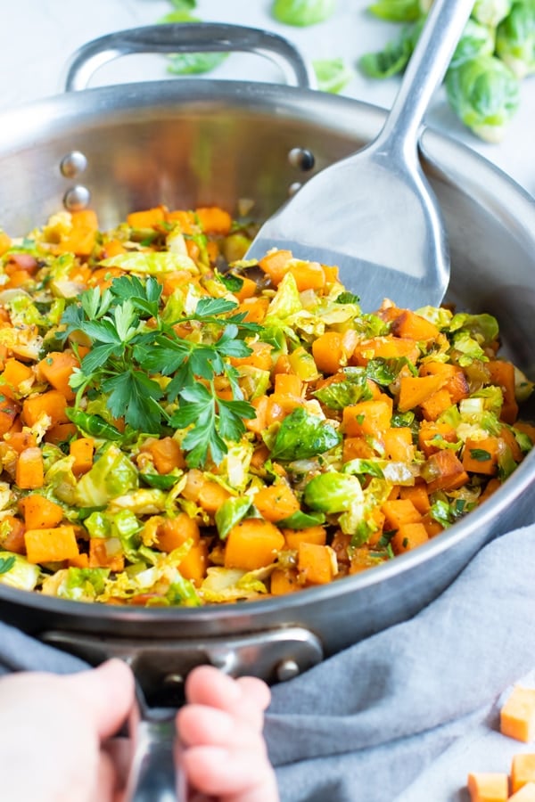 A hand holding a skillet of gluten-free Brussel sprouts and sweet potatoes with a silver spatula inside.