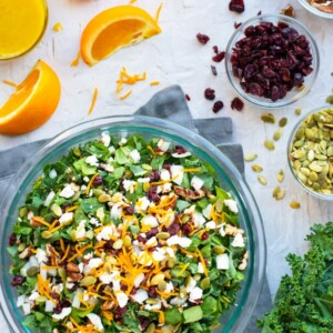 Thanksgiving salad recipe in a clear bowl with oranges, cranberries, and pumpkin seeds surrounding it.