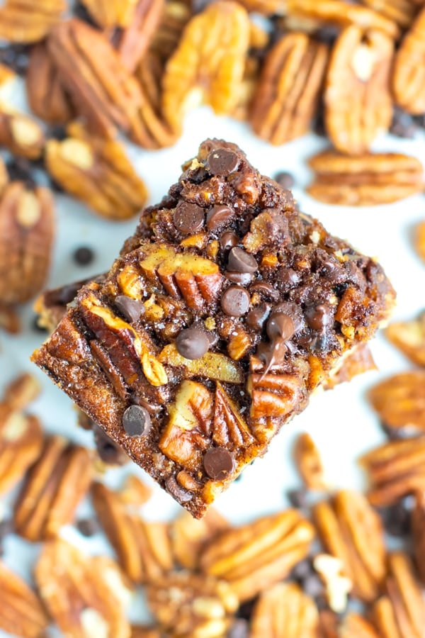 Chocolate pecan pie bars in a stack with pecans and chocolate chips surround them.