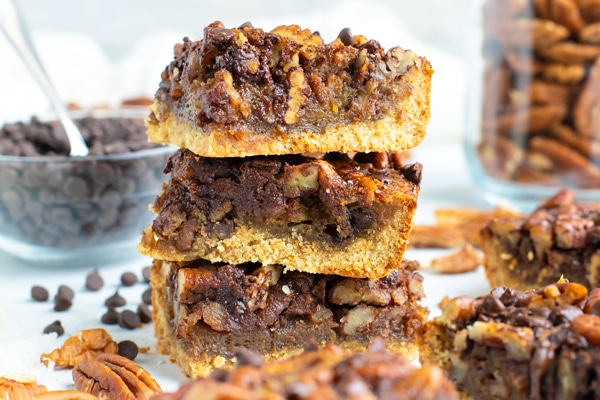 An easy pecan pie bars recipe in a stack with chocolate chips on a white background.