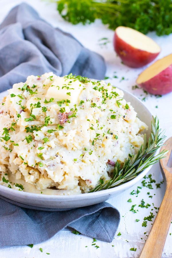 Garlic mashed potatoes in a white bowl with a wooden spoon and red potatoes.
