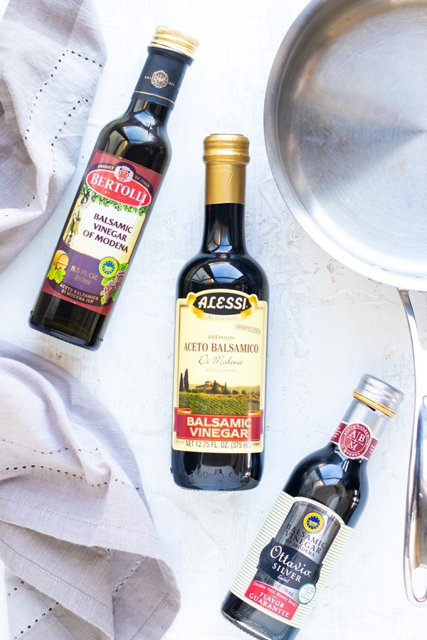 Different brands of balsamic vinegar of the Modena region on a white surface with a stainless steel saucepan.