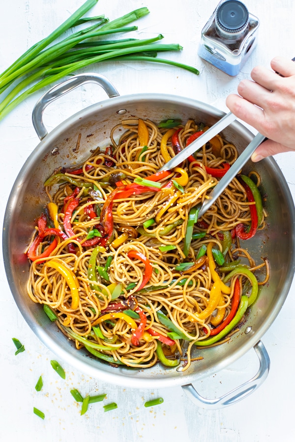 A hand with tongs tossing around a wok full of sesame noodles with garlic, ginger, and bell peppers.