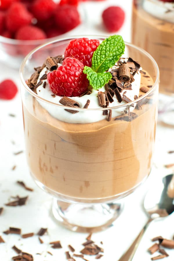 A dessert glass full of chocolate mousse topped with whipped coconut cream and garnished with chocolate curls, raspberries, and mint leaves.