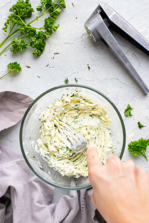 A hand mixing together garlic herb butter in a clear bowl.