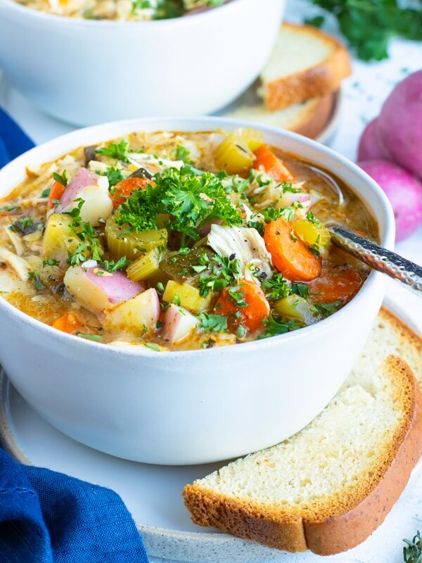 A white bowl full of Instant Pot chicken vegetable soup with two pieces of bread and red potatoes next to it.