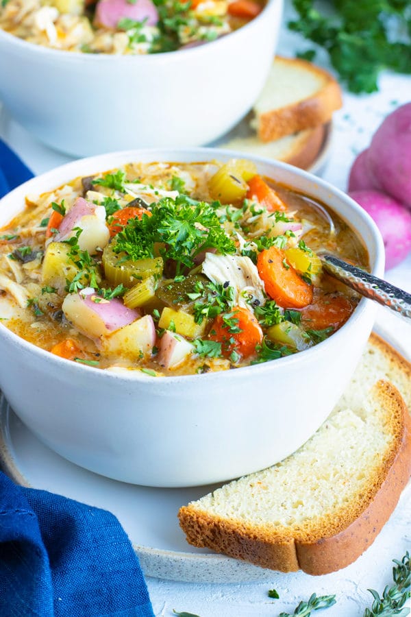 A white bowl full of Instant Pot chicken vegetable soup with two pieces of bread and red potatoes next to it.