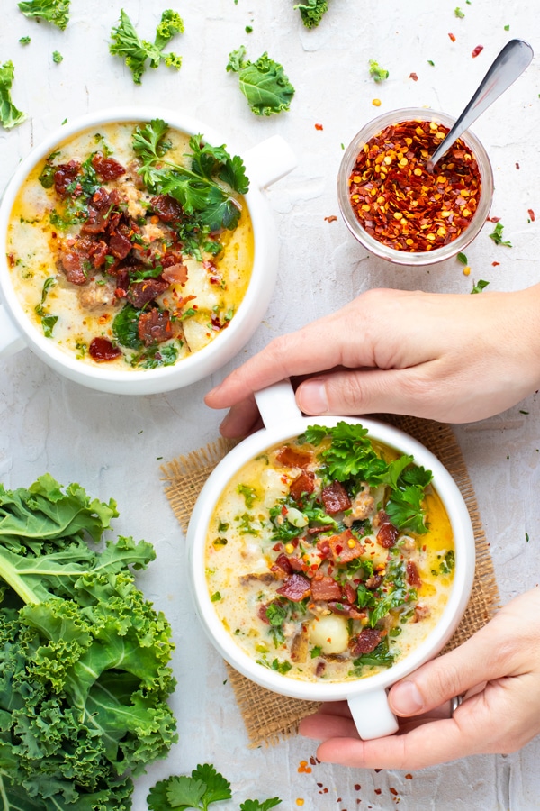 Two white bowls full of a zuppa toscana soup recipe next to kale and red pepper flakes.