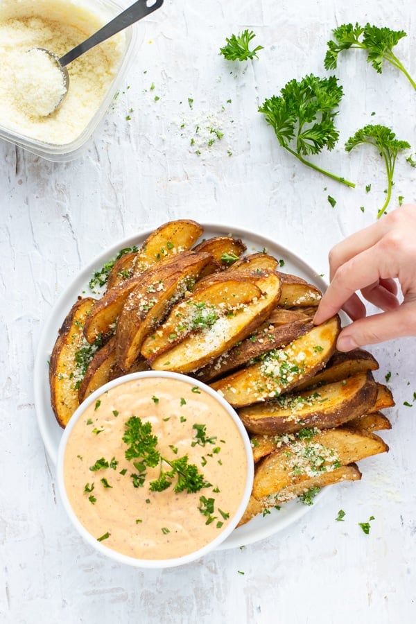 A hand grabbing a roasted potato wedge from a plate of them.