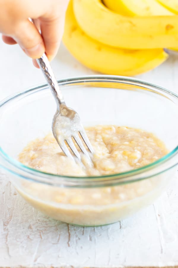 A clear bowl with ripe bananas being mashed by a fork.