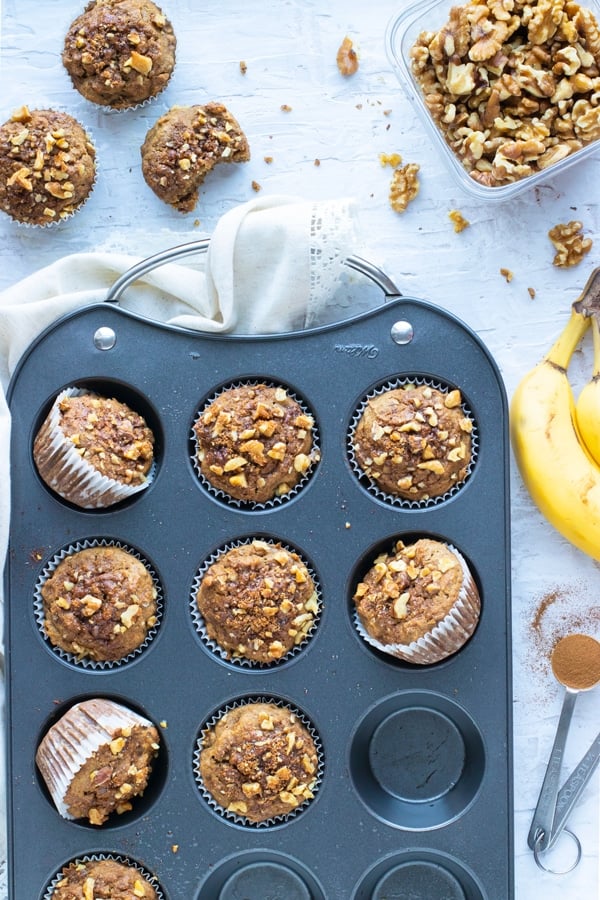 A muffin tray full of healthy banana bread muffins next to a tub of walnuts and two bananas.