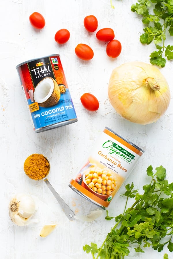 A can of coconut milk, garbanzo beans, and a sweet onion next to cherry tomatoes and cilantro.