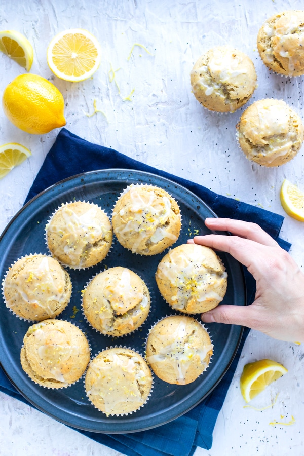 A navy blue plate full of eight gluten-free lemon poppy seed muffins and a hand grabbing one of them.