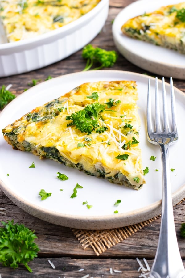 A white serving plate with a slice of spinach crustless quiche and a large baking dish full of it.