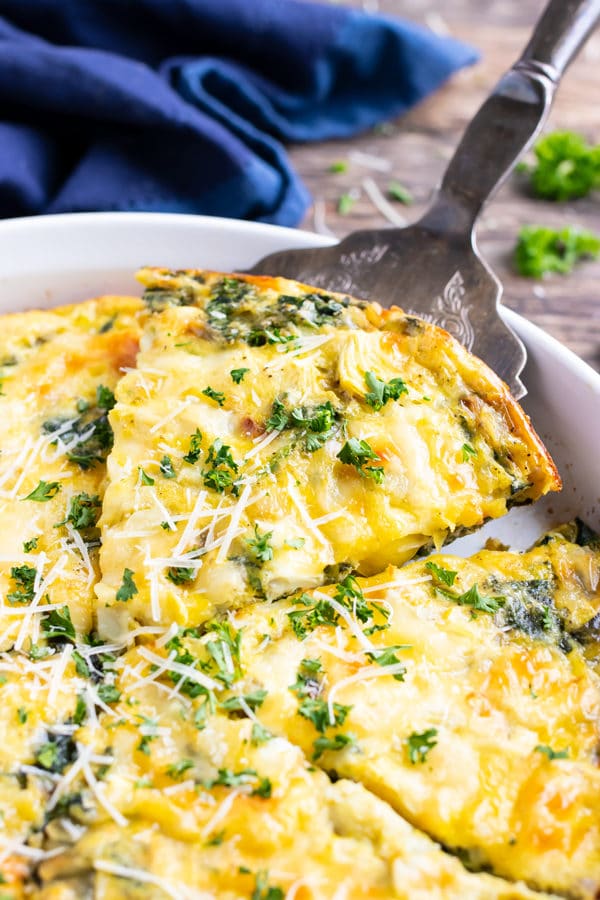 A slice of an egg bake recipe with spinach and cheese being picked up out of a casserole dish.