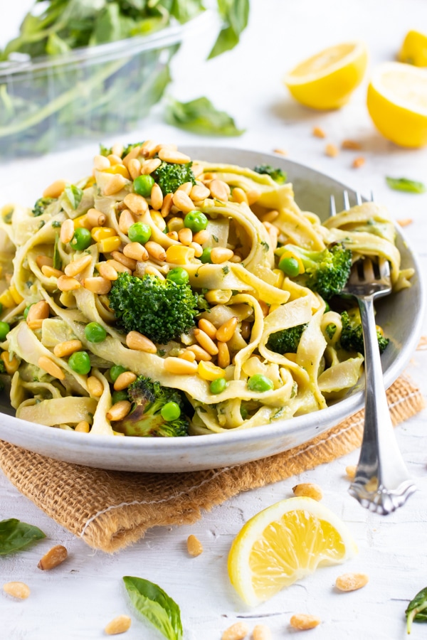 A white bowl full of avocado pesto pasta with broccoli, pine nuts, and corn.