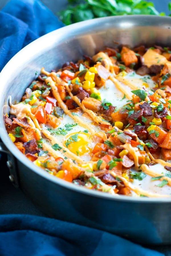 A stainless steel skillet full of sweet potato hash browns with eggs and sriracha mayo.