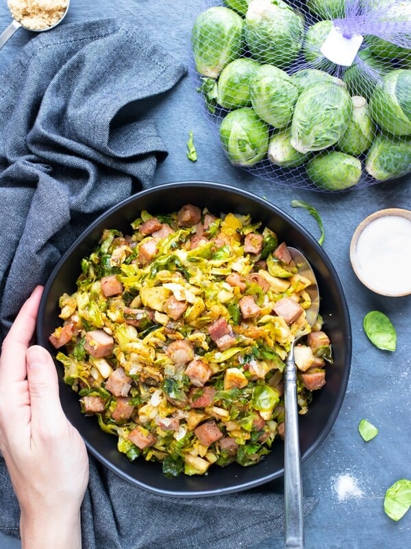 A bag full of Brussels sprouts next to a serving bowl full of sauteed and shredded Brussels sprouts with cubed ham.