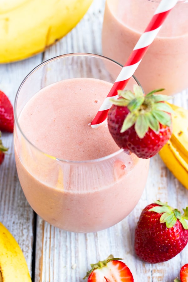 A glass full of a strawberry and banana smoothie with a strawberry as a garnish and a red/white striped straw.