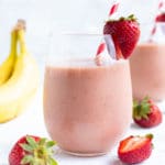 Two clear glasses full of a strawberry banana smoothie recipe with a red and white striped straw.