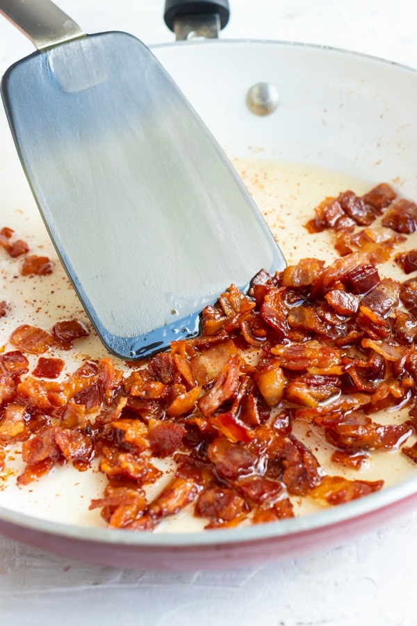 Bacon being cooked in a ceramic skillet with a spatula stirring it.