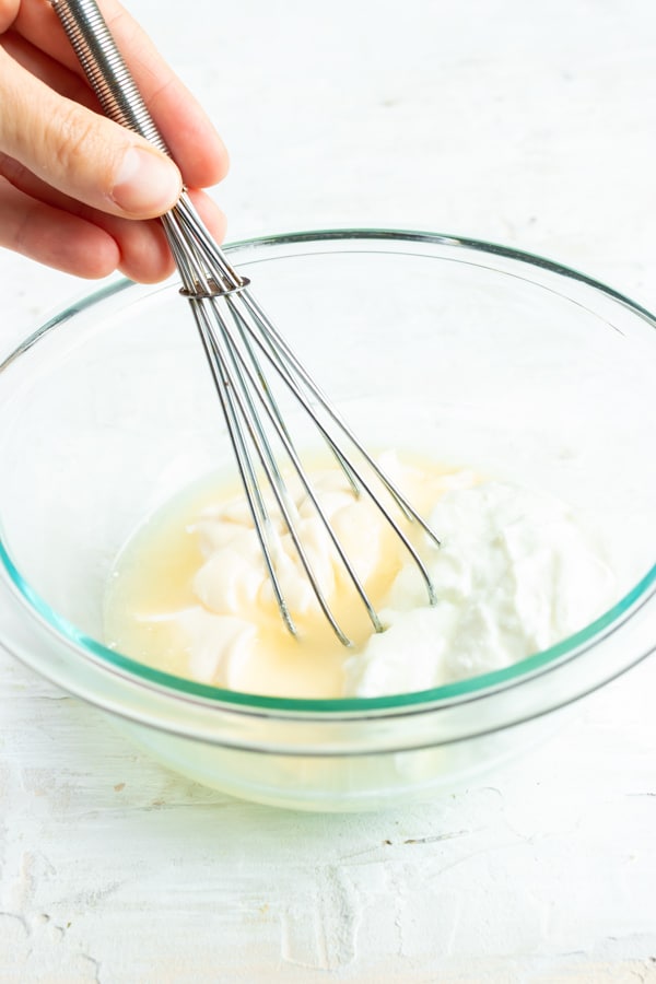 A hand whisking together a broccoli salad dressing made from mayonnaise, white wine vinegar, Greek yogurt, and sugar.