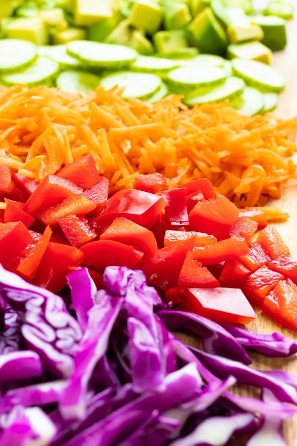 Purple cabbage, red bell pepper, grated carrots, cucumber, and avocado on a cutting board.