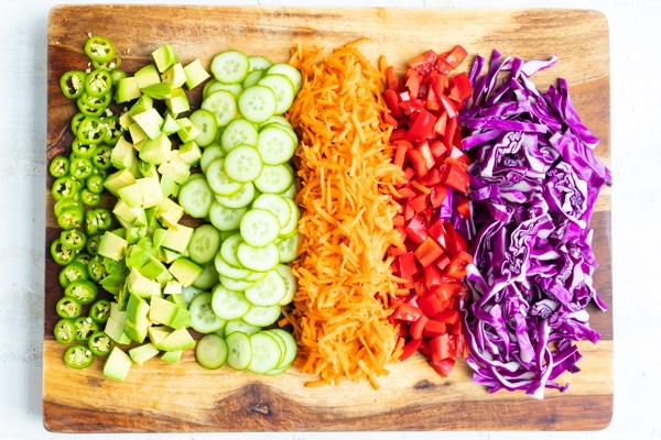 A cutting board with chopped jalapeno, purple cabbage, red bell pepper, grated carrots, cucumber, and avocado to go in a vegan sushi bowl recipe.