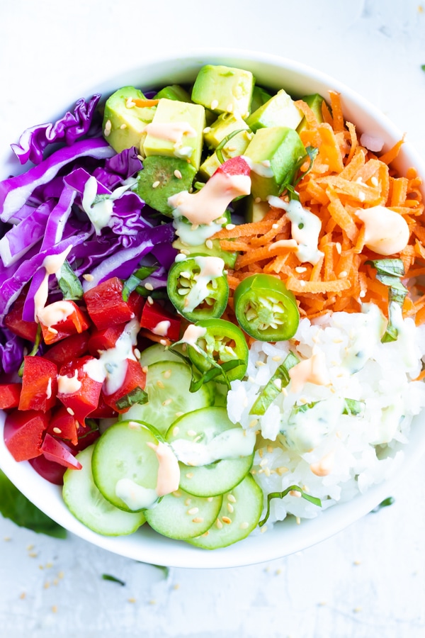 Deconstructed sushi bowl recipe with avocado, cabbage, carrots, cucumber and sauce drizzled on top.