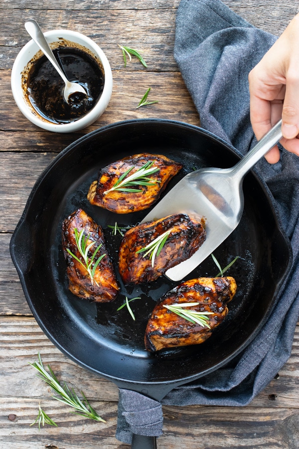 Honey balsamic chicken breast being picked up from a skillet with a spatula with a bowl of balsamic glaze next to it.