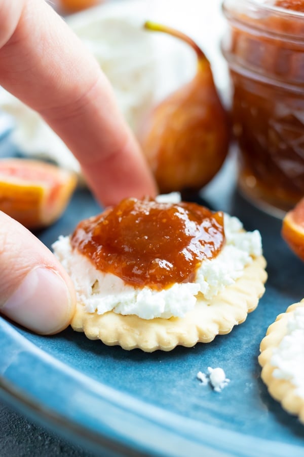 A hand picking up a cracker with goat cheese and jam.