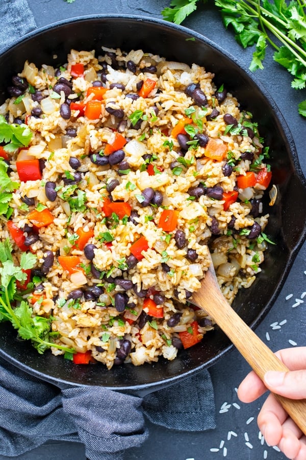 A hand scooping out some gallo pinto from a cast-iron skillet with a wooden spoon.