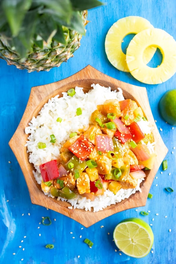 Easy pineapple chicken recipe served with white rice in a wooden bowl next to pineapple rings and limes.