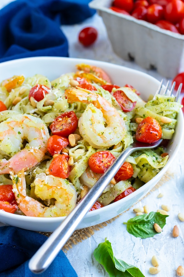 A bowl full of shrimp pesto pasta with tomatoes, Parmesan cheese, and pine nuts with a carton of cherry tomatoes behind it.