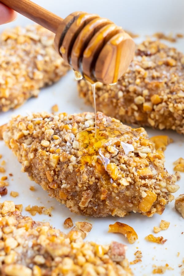 Honey being drizzled over baked pecan crusted chicken with a gluten-free pecan crust in a baking dish.