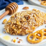 A healthy gluten-free honey baked chicken breast recipe on a white dinner plate next to pretzels and pecans.