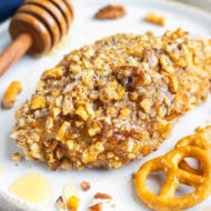 A healthy gluten-free honey baked chicken breast recipe on a white dinner plate next to pretzels and pecans.